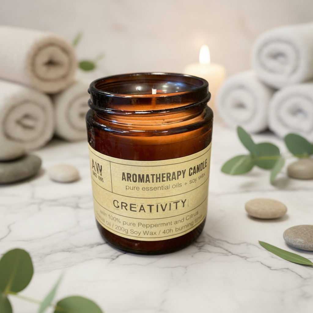 A soy wax candle with a label that reads 'AROMATHERAPY CANDLE creativity', burning inside a clear glass jar on a white coaster, with green leaves in the background.