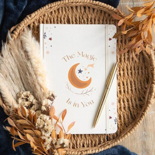 Notebook with 'The Magic Is in You' design on a woven mat with dried leaves and a pen.