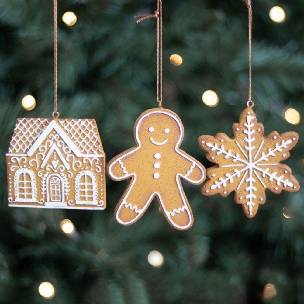 Three Christmas ornaments shaped like a gingerbread house, gingerbread man, and snowflake hanging against a blurred Christmas tree background.
