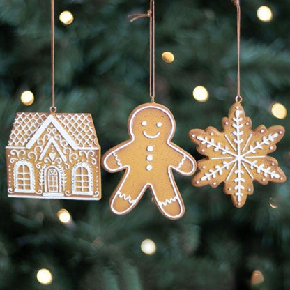 Three Christmas ornaments shaped like a gingerbread house, gingerbread man, and snowflake hanging against a blurred Christmas tree background.