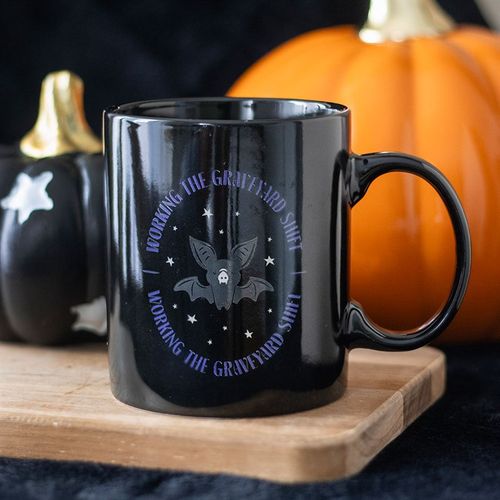 Black mug with a bat design and text on a wooden surface with pumpkins in the background
