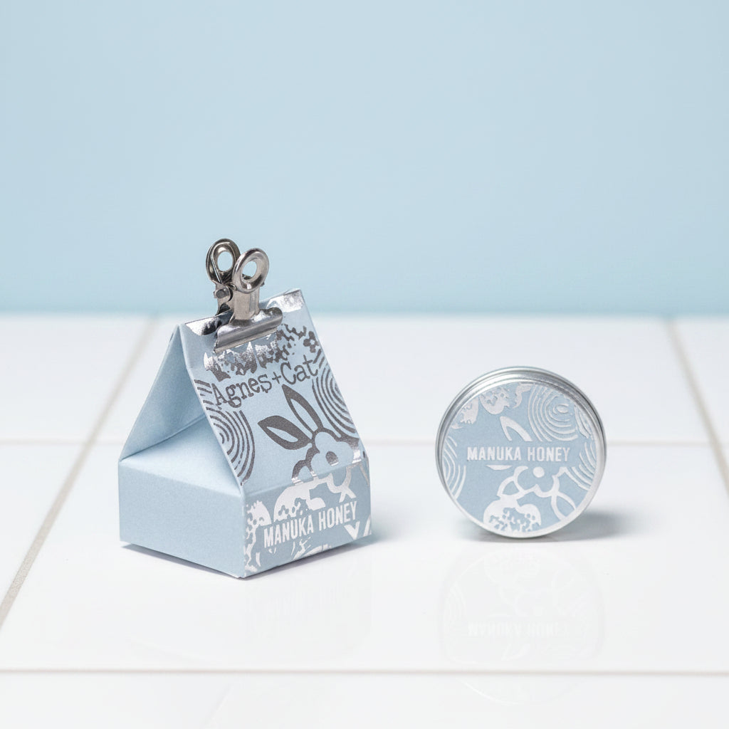 A triangular pale blue packaging of Agnes and Cat lip balm with a clip on top and 'manuka Honey' text on the front.