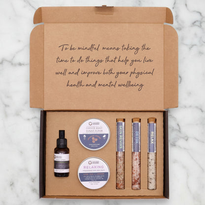 A self-care gift set box containing various bath products including a bottle, possibly of essential oil or perfume, and tubes with scrub and lotion.