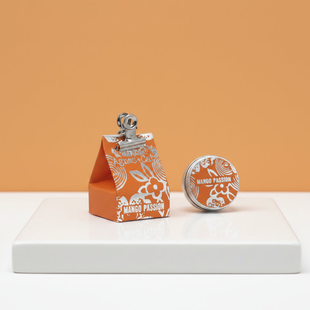 A triangular orange packaging of Agnes and Cat lip balm with a clip on top and 'mango passion' text on the front.