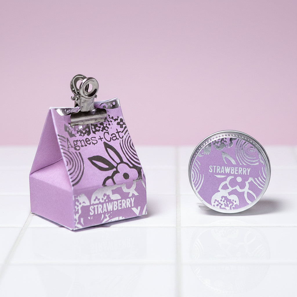 A triangular purple packaging of Agnes and Cat lip balm with a clip on top and 'strawberry' text on the front.